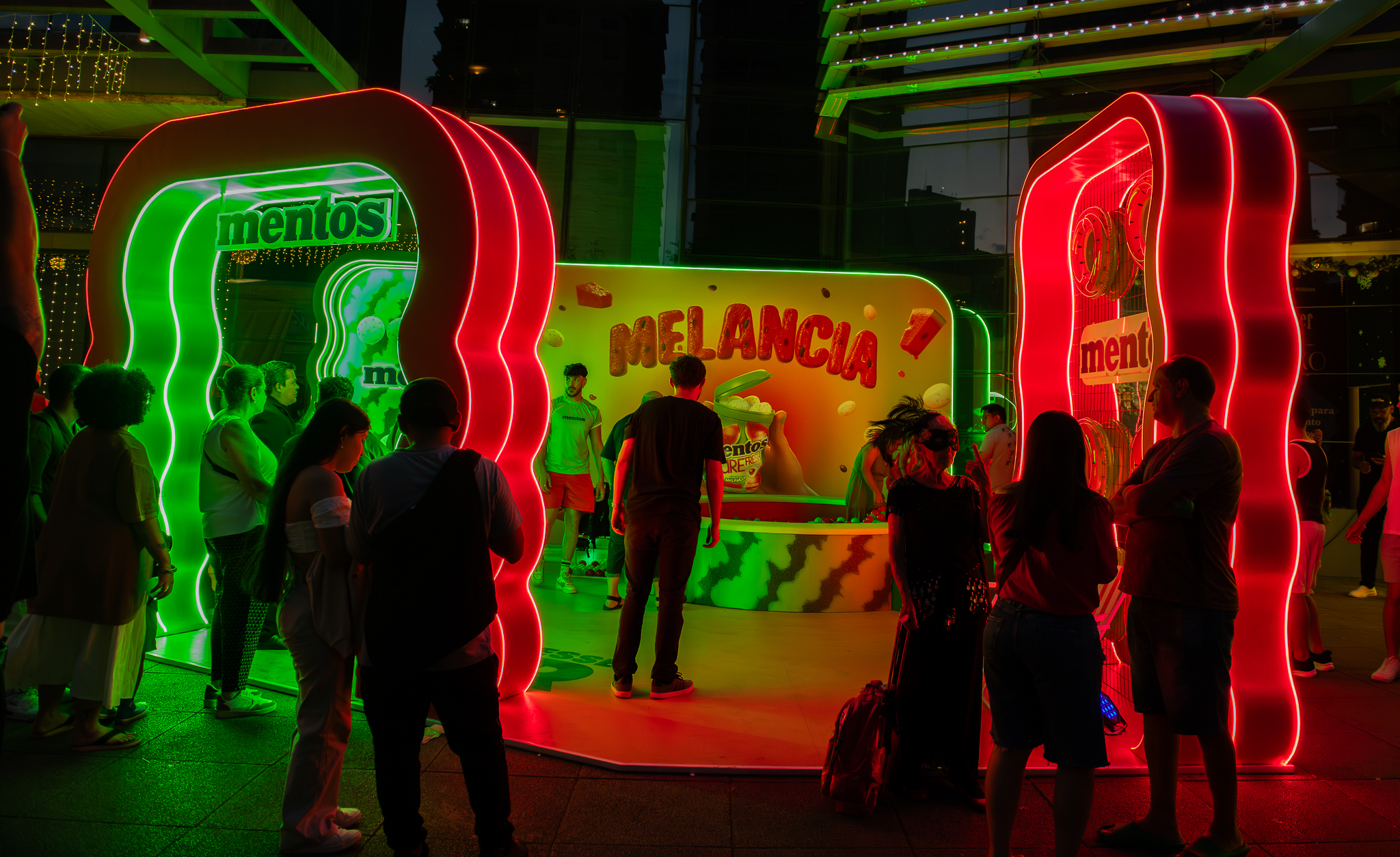 Instalação neon verde e vermelho da Mentos Melancia com público em ativação noturna em shopping center.